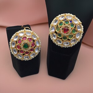 Puede incluir: Dos piezas de joyer&iacute;a redondas de tono dorado con detalles de perlas blancas y piedras preciosas de colores. Las piezas presentan una piedra preciosa verde central rodeada de piedras preciosas verdes, rojas y blancas m&aacute;s peque&ntilde;as.