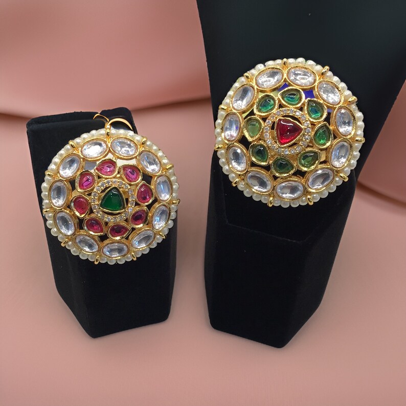 Puede incluir: Dos piezas de joyer&iacute;a redondas de tono dorado con detalles de perlas blancas y piedras preciosas de colores. Las piezas presentan una piedra preciosa verde central rodeada de piedras preciosas verdes, rojas y blancas m&aacute;s peque&ntilde;as.