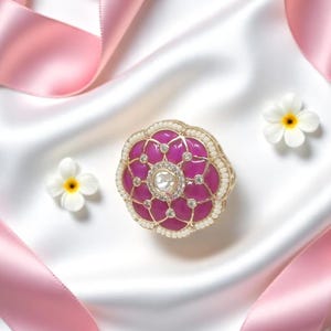 May include: A floral-shaped jewelry box with a magenta enamel finish, adorned with clear gemstones and a central diamond. The box is set on a white satin surface, with pink ribbons and small white flowers with yellow centers.