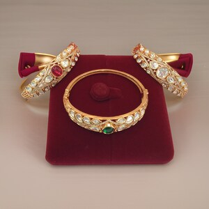 May include: Three gold bangles with intricate designs featuring white and red gemstones. The bangles are displayed on a red velvet cushion.