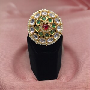 Puede incluir: Un anillo de oro con un dise&ntilde;o redondo con piedras preciosas blancas, verdes y rojas. El anillo est&aacute; colocado sobre un soporte de terciopelo negro.