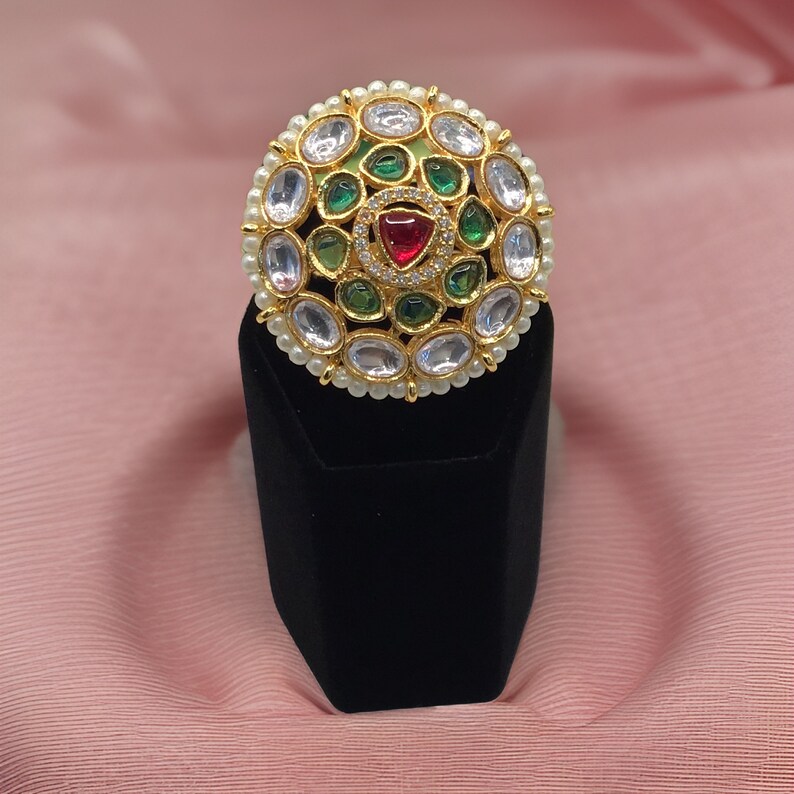 Puede incluir: Un anillo de oro con un dise&ntilde;o redondo con piedras preciosas blancas, verdes y rojas. El anillo est&aacute; colocado sobre un soporte de terciopelo negro.