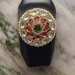 Puede incluir: Un anillo de oro con un dise&ntilde;o circular que presenta piedras preciosas blancas, rosas y verdes. El anillo est&aacute; colocado en un expositor negro.