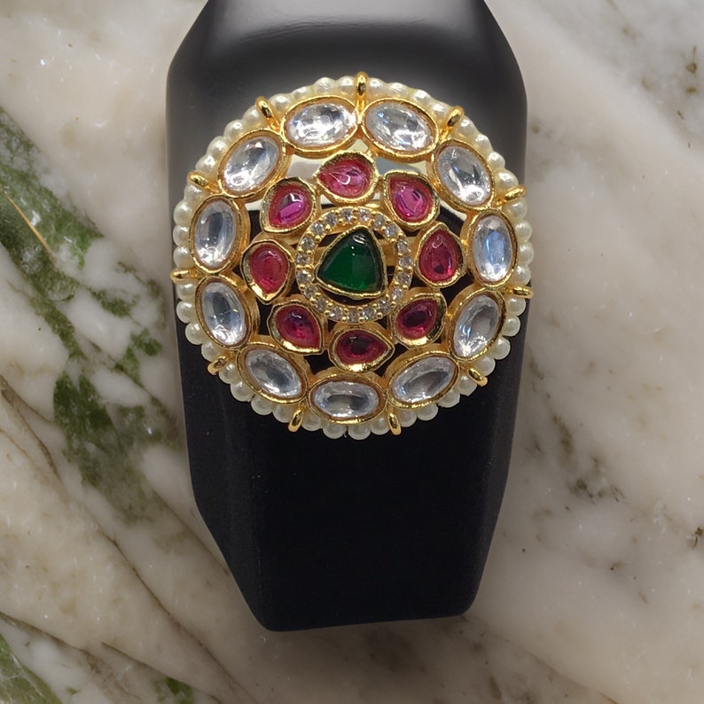 Puede incluir: Un anillo de oro con un dise&ntilde;o circular que presenta piedras preciosas blancas, rosas y verdes. El anillo est&aacute; colocado en un expositor negro.