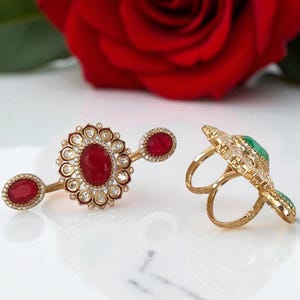 May include: Two gold-toned rings with ornate designs. One ring features a large oval red gemstone surrounded by smaller clear stones. The other ring has a green gemstone. A red rose is in the background.