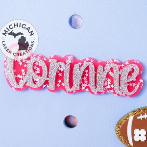 May include: Pink glitter name "Brinne" with silver glitter stars on a light blue background. A small brown football with gold trim and the number "72" is in the lower right corner. A round sticker with the text "Michigan Laser Creations" is in the upper left corner.