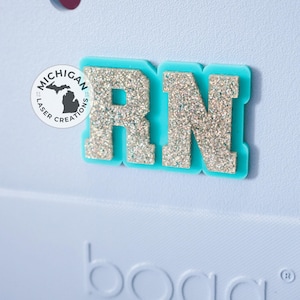 May include: A teal and glitter RN badge. The letters are silver glitter on a teal background. The badge is on a light blue surface. The Michigan Laser Creations logo is in the upper left corner.