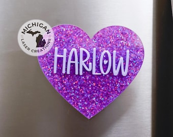 May include: Purple glitter heart with the name Harlow written in white letters. The heart is on a silver surface. The image also includes a small logo with the text "Michigan Laser Creations".