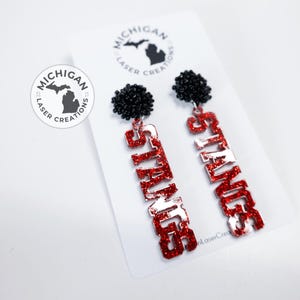 May include: A pair of red and white glitter earrings with black pom-poms. The earrings spell out "SPARTANS".