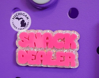Könnte beinhalten: Ein pinkfarbener "SNACK DEALER"-Anhänger mit Glitzerakzenten, vor einem lila Hintergrund. Der Anhänger ist ein Schuhzubehör. Ein Logo von Michigan Laser Creations ist ebenfalls sichtbar.