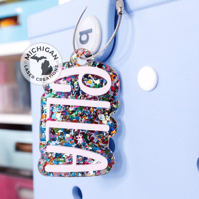 May include: A colorful, glittery keychain with the name "Aila" written in white letters. The keychain is attached to a silver chain and has a small, round, blue tag with the letter "b" on it. The keychain is made by "Michigan Laser Creations".