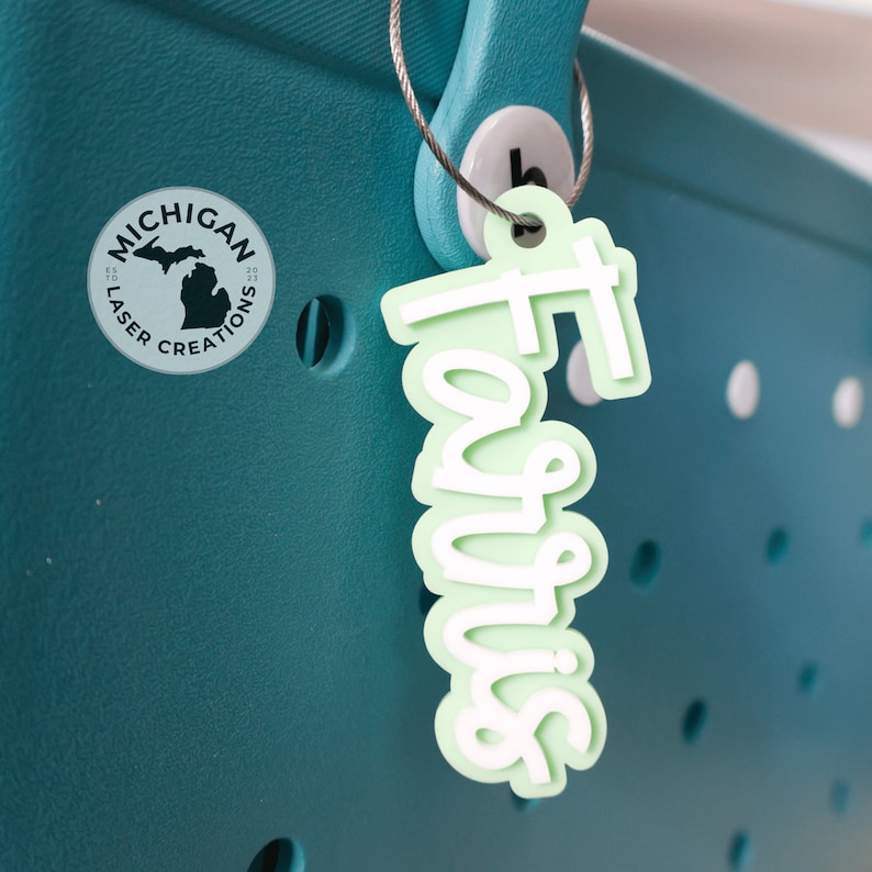 May include: A teal blue tote bag with a white circle logo that says "Michigan Laser Creations" and a light green charm that says "Favors" hanging from a silver chain.