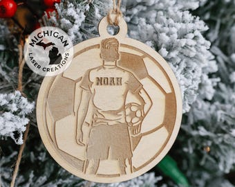 May include: A wooden ornament featuring a silhouette of a soccer player holding a ball. The ornament is round and has a brown wood finish. The player's jersey has the name "Noah" printed on the back. The ornament is decorated with a soccer ball pattern.
