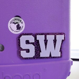 May include: A purple item with a white "SW" letter design outlined in purple glitter. A circular sticker with "Michigan Laser Creations" and a Michigan outline is also visible. The item is a solid purple color.