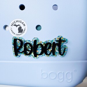May include: A blue and white personalized name tag with the name "Robert" in black script lettering. The name tag is decorated with blue and gold glitter. The tag is on a light blue background with a white circle that says "Michigan Laser Creations".