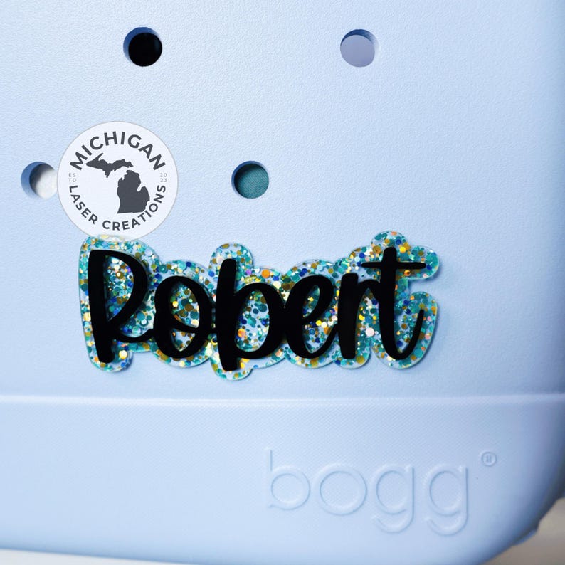 May include: A blue and white personalized name tag with the name "Robert" in black script lettering. The name tag is decorated with blue and gold glitter. The tag is on a light blue background with a white circle that says "Michigan Laser Creations".