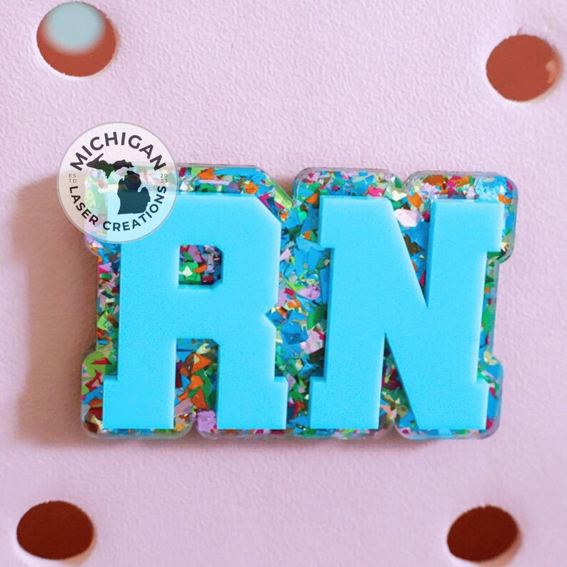 May include: A light blue RN badge with a colorful confetti border. The letters are bold and stand out against the confetti background. The badge has a Michigan Laser Creations logo on the top left corner. The background is a light pink.