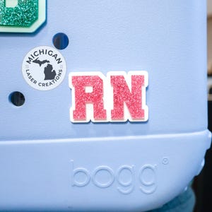 May include: A light blue tote bag with a pink glitter "RN" patch. A round sticker with the outline of Michigan and the words "Michigan Laser Creations" is also visible. The bag has the word "bogg" embossed on the front.