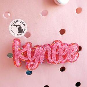 May include: Pink glitter name "Kynlee" with a Michigan Laser Creations logo in the background.