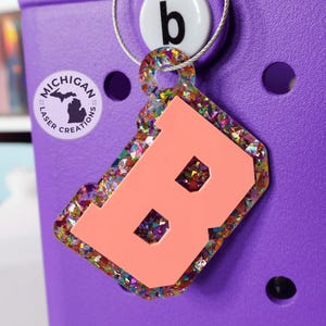 May include: A pink letter "B" with a white outline, surrounded by colorful glitter, on a purple background. The letter is attached to a silver ring with a small white circle with the letter "b" on it. The letter is a charm or keychain with the text "Michigan Laser Creations" on a small sticker.