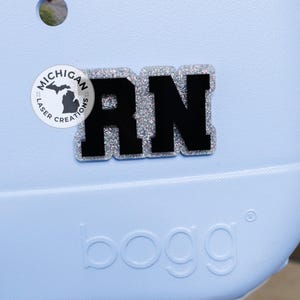 May include: A light blue bag with a black and silver glitter RN charm. The charm is a Registered Nurse initial. The bag has the word "bogg" printed on the front and a Michigan Laser Creations logo.
