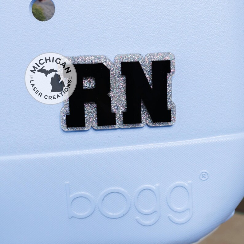 May include: A light blue bag with a black and silver glitter RN charm. The charm is a Registered Nurse initial. The bag has the word "bogg" printed on the front and a Michigan Laser Creations logo.