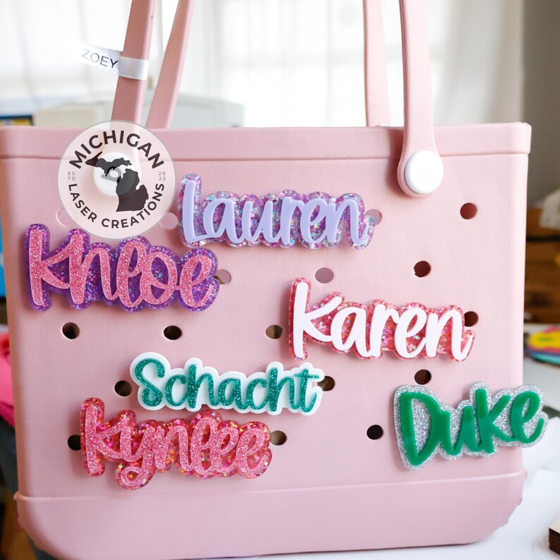May include: A pink tote bag with a variety of personalized name tags. The name tags are made of glitter and have a white outline. The names on the tags are "Lauren", "Knobe", "Karen", "Schacht", "Kynlee", and "Duke".