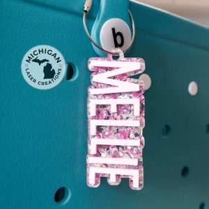 May include: A pink and white glitter charm with the name "Millie" written in white letters. The charm is attached to a silver chain and is hanging from a teal tote bag.