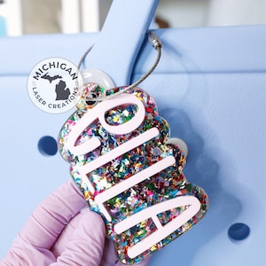 May include: A white personalized name tag with the name "Alia" in white letters. The tag is filled with colorful confetti and attached to a silver cable.