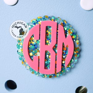 May include: Pink monogram initial "CBM" with a glittery, colorful background. The background is a mix of blue, green, yellow, and white glitter.