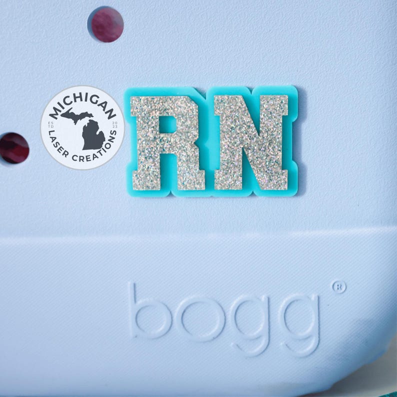 May include: A light blue bag with a glittery silver "RN" charm. The charm is outlined in a matching light blue. A round sticker with the text "Michigan Laser Creations" and a silhouette of the state is also visible.