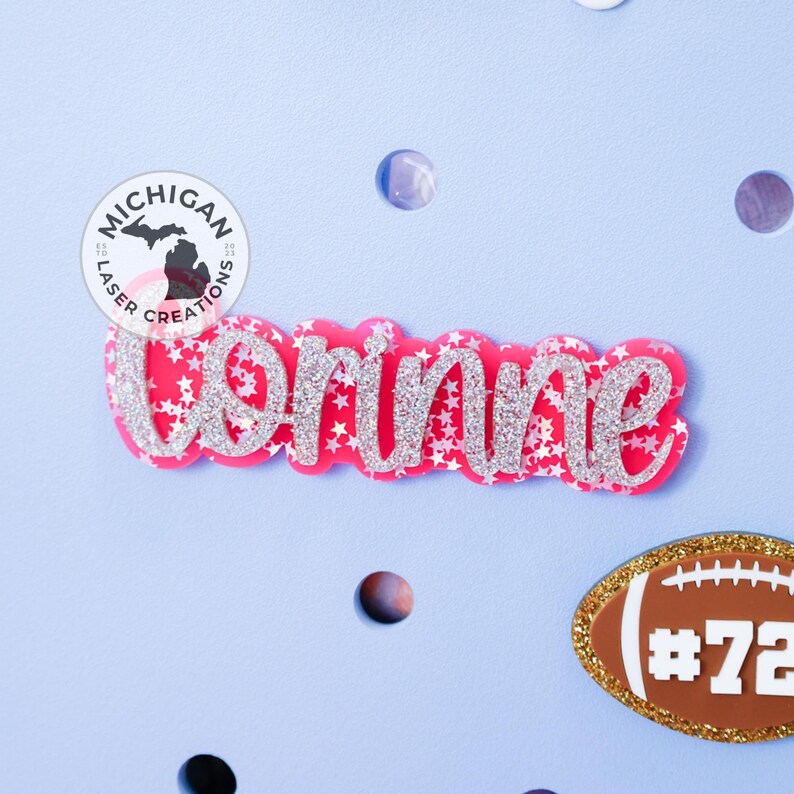 May include: Pink glitter name "Brinne" with silver glitter stars on a light blue background. A small brown football with gold trim and the number "72" is in the lower right corner. A round sticker with the text "Michigan Laser Creations" is in the upper left corner.