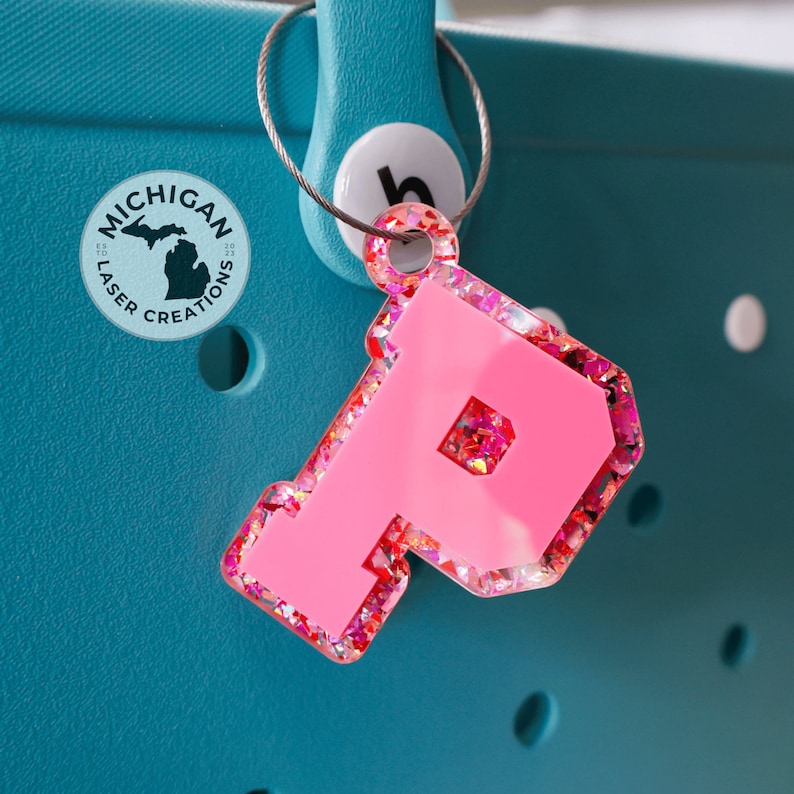 May include: Pink glitter letter P charm with a white background. The charm is attached to a silver key ring. The charm is hanging from a teal blue tote bag with a white circle that says "Michigan Laser Creations" on it.