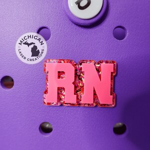 May include: A close-up of a purple shoe with a pink and glittery "RN" charm. A round charm with the text "Michigan Laser Creations" and a state outline is also visible. Additional white and black charms are also visible.