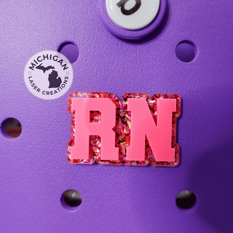 May include: A close-up of a purple shoe with a pink and glittery "RN" charm. A round charm with the text "Michigan Laser Creations" and a state outline is also visible. Additional white and black charms are also visible.