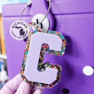 May include: A white letter C with a purple outline, covered in colorful confetti, attached to a silver key chain with a round tag that says "Michigan Laser Creations" in black text.