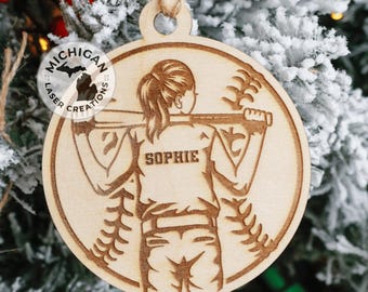 May include: A wooden Christmas ornament featuring a silhouette of a person holding a baseball bat. The ornament is round and has a brown wood grain. The person is wearing a baseball uniform and has the name "Sophie" written on the back. The ornament is decorated with a baseball stitch pattern.