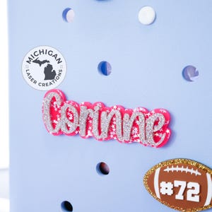 May include: A light blue wall with a pink glitter name "Corinne" and a brown football with the number "#72" on it. There is a white circle with a black outline and the text "MICHIGAN LASER CREATIONS" and a black outline of the state of Michigan.