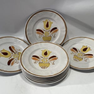 Puede incluir: Un juego de platos de cerámica con un fondo blanco roto moteado y un borde marrón. Cada plato presenta un diseño floral con flores amarillas y marrones en forma de tulipán en una cesta amarilla. El juego incluye platos de diferentes tamaños y cantidades.