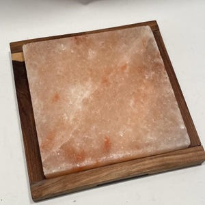Puede incluir: Un bloque de sal del Himalaya cuadrado, de unos 20 cm x 20 cm, de color rosa anaranjado, colocado en una bandeja de madera oscura. El bloque de sal tiene una superficie texturizada. La bandeja de madera tiene vetas naturales.