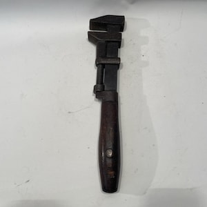 Puede incluir: Una llave ajustable con una cabeza de metal oscuro y un mango de madera. La llave tiene un mecanismo de tornillo para ajustar el tamaño de la mordaza. El mango es de color marrón oscuro y tiene forma redondeada.