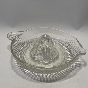 May include: A clear glass citrus juicer with a ribbed bowl and a central reamer. The juicer has a pouring spout and a handle. The design is simple and functional, suitable for kitchen use.