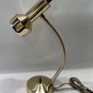 Puede incluir: Lámpara de escritorio vintage con acabado dorado. La lámpara presenta un brazo flexible de cuello de cisne, una base redonda y una pantalla cónica. La parte superior de la lámpara tiene un acento de madera. La lámpara está diseñada para uso en escritorio.
