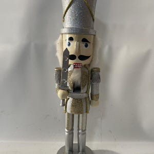Puede incluir: Una figura de cascanueces plateada y dorada con un sombrero alto y brillante y una expresión severa. La figura sostiene una espada plateada y tiene barba y bigote blancos. La base es plateada y redonda, y el uniforme tiene detalles dorados.