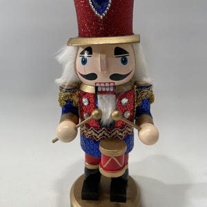 Peut inclure: Un casse-noisette décoratif en bois avec un chapeau haut de forme rouge à paillettes et un bijou bleu. Le casse-noisette a une perruque blanche, un visage peint et porte un uniforme rouge, bleu et or. Il tient des baguettes et un petit tambour rouge.
