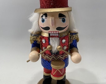 May include: A decorative wooden nutcracker soldier in a red glitter top hat with a blue jewel. The nutcracker has a white wig, a painted face, and is dressed in a red, blue, and gold uniform. It holds drumsticks and a small red drum.