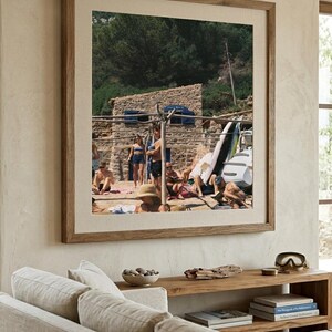 May include: A framed photograph of a beach scene with people, a stone building, and a green hillside. The photograph is in a light brown wooden frame and hangs above a wooden console table.
