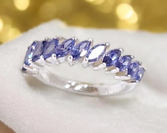 May include: A silver ring featuring a row of marquise-cut, light blue gemstones. The gemstones are set closely together, creating a continuous line of color. The ring is displayed on a white surface with a blurred, golden background.