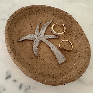 May include: A brown ceramic dish with a white palm tree design. The dish has two gold rings with clear stones on it.