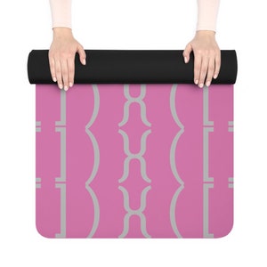 May include: A rolled-up yoga mat with a black top and a pink surface. The pink surface features a repeating gray geometric pattern. The mat is held by two hands with light skin.